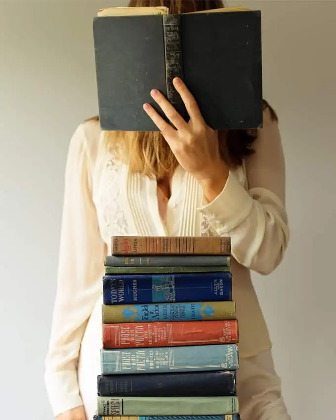 mehrere Bücher liegen übereinander, eine Frau nimmt ein Buch und schlägt es vor ihrem Gesicht auf