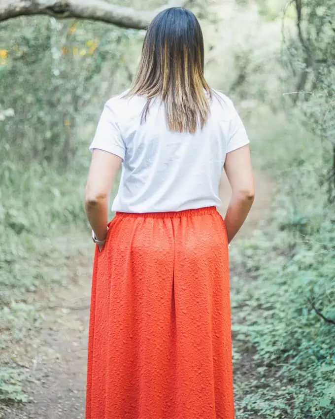 Bild einer Frau von hinten. Sie trägt ein weißes Hemd und einen langen orangefarbenen Rock