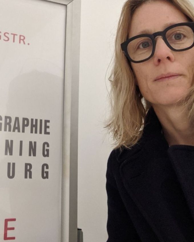 Frau mit blonden schulterlangen Haaren und schwarzer Brille und schwarzem Mantel vor einem Schild zur Mammographie.