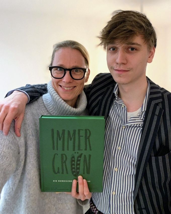 Frau im Rollenkragen Pullover und Mann im Sakko und Hemd. Beide halten ein grünes Kochbuch in der Hand.