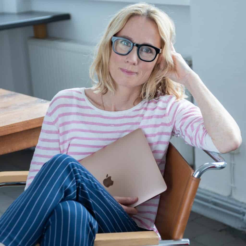 Gründerin Susanne Liedtke von nobodytoldme.com sitz in einem Stuhl in einer Küche, lächelt in die Kamera und hält ihr Macbook in der Hand.