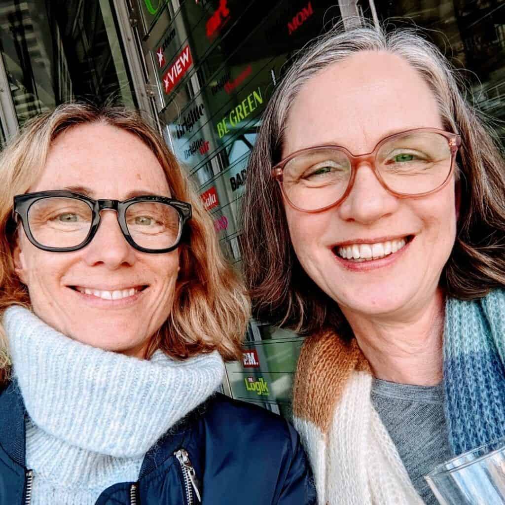 zwei Frauen mittleren Alters, die beide eine Brille tragen, lächeln in die Kamera. Sie stehen draußen vor dem Eingang des Gruner & Jahr Gebäudes in Hamburg