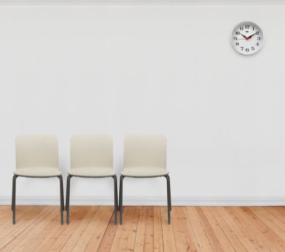drei weiße Stühle mit schwarzen Beinen aus Metall stehen in einem weißen Raum auf hellem Kiefernparkett. An der Wand oben rechts hängt eine schlichte runde mit schwarzen Zeigern und Zahlen. Es ist ein Warteraum bei der Ärztin.