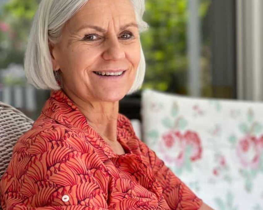 65 j&auml;hrige Frau mit wei&szlig;em Bob l&auml;chelt in die Kamera. Sie tr&auml;gt ein Koralle farbenes Kleid und sitzt auf einem Rattanstuhl.