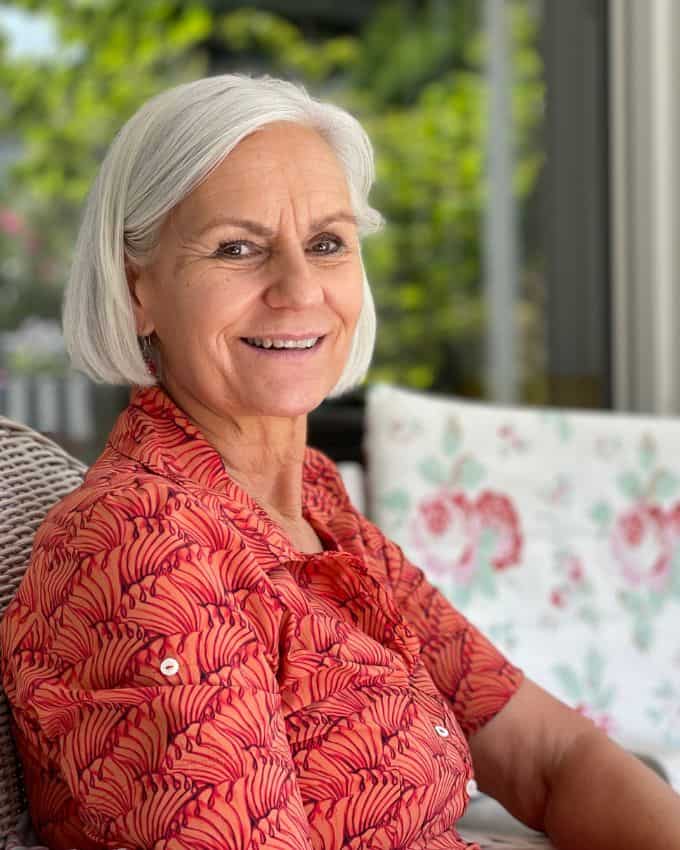 65 jährige Frau mit weißem Bob lächelt in die Kamera. Sie trägt ein Koralle farbenes Kleid und sitzt auf einem Rattanstuhl.