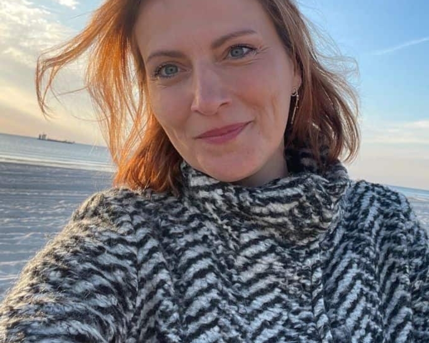 Frau 40+ (2) 42 jährige Frau lächelt zufrieden in die Kamera. Sie trägt einen Sonnenbrille aus dem Kopf und hat rot-braune Haare. Sie trägt einen schwarz-weiß gemusterten kuscheligen Pulli. Im Hintergrund ist der Strand und das Meer zu sehen.