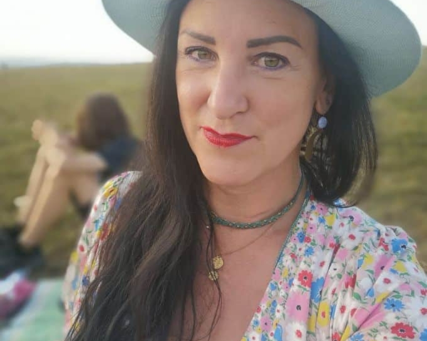 Frau mit braunen langen Haaren, trägt einen hellblauen Hut und ein ausgeschnittenes Sommerkleid mit Blumenprint. Um den Hals trägt sie zwei kurze Ketten. Sie steht am Deich und ist 50 Jahre alt.