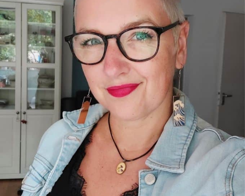 Frau mit wasserstoffblondem Kurzhaarschnitt und pinkem Lippenstift schaut zuversichtlich in die Kamera; sie trägt auffällige Hänge-Ohrringe