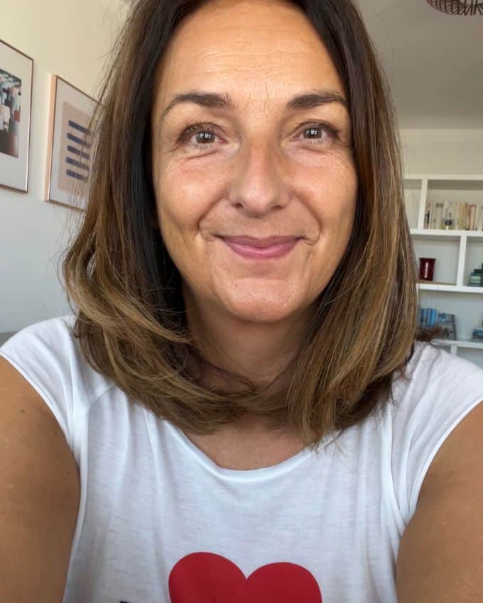 Selfie einer sehr zufriedenen Frau mit weißem T-Shirt mit Herz Print. Sie trägt einen braunen, langen Bob Haarschnitt. Ihre Haut ist leicht gebräunt. Sie steht in ihrem Wohnzimmer.
