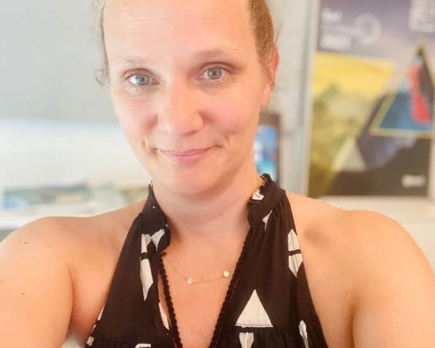 Frauen mit blonden Haaren, die zurückgebunden sind, trägt ein schwarzes Neckholderoberteil, auf dem wei&szlig;e Dreiecke zu sehen sind. Es ist Sommer. Sie ist 44.