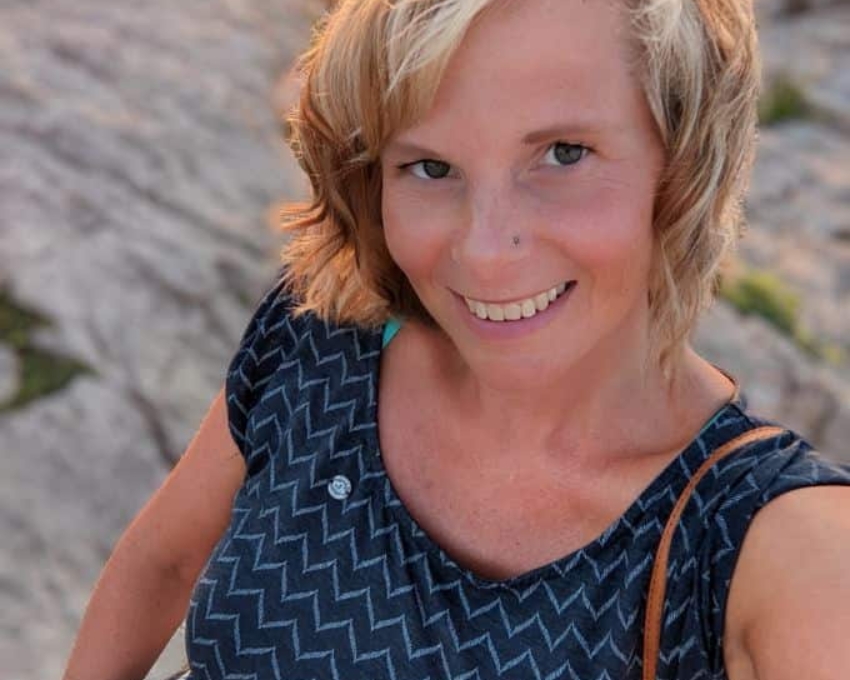 So sieht 41 aus Frau mit blondem Bob steht auf felsigem Grund. Sie trägt ein dunkleblaues körperbetontes T-Shirt und trägt über die Schulter den Lederriemen einer Handtasche. Am rechten Arm trägt sie zwei Armbänder. Eines davon ist gelb.