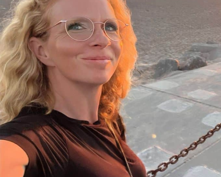 So sieht 43 aus. 43 jährige Frau, die am Strand bei Sonnenuntergang ein Selfie von sich macht. Sie hat rotblonde Haare, trägt eine goldene, runde Brille und ein schwarzes T-Shirt.