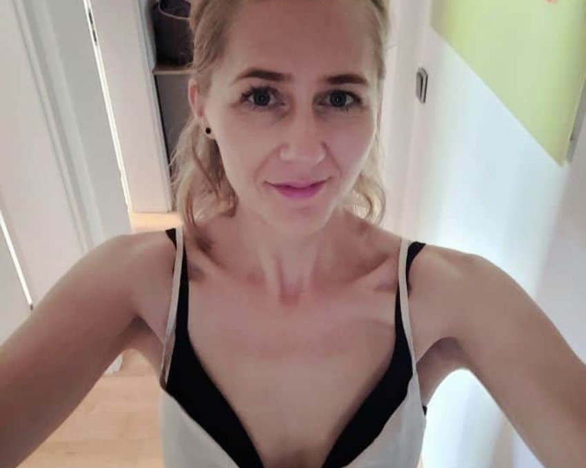So sieht 45 aus Eine Frau mit schwarzem BH und weißem Träger Top macht ein Selfie von sich im Flur. Sie ist 45 und trägt ihr dunkelblondes Haar zurückgesteckt.