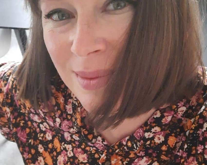 So sieht 48 aus 48 jährige Frau mit braunem glattem Bob und Pony schaut zufrieden in die Kamera. Sie trägt eine Bluse mit Blumenmuster in braun, orange, rosa und dunkelrosa