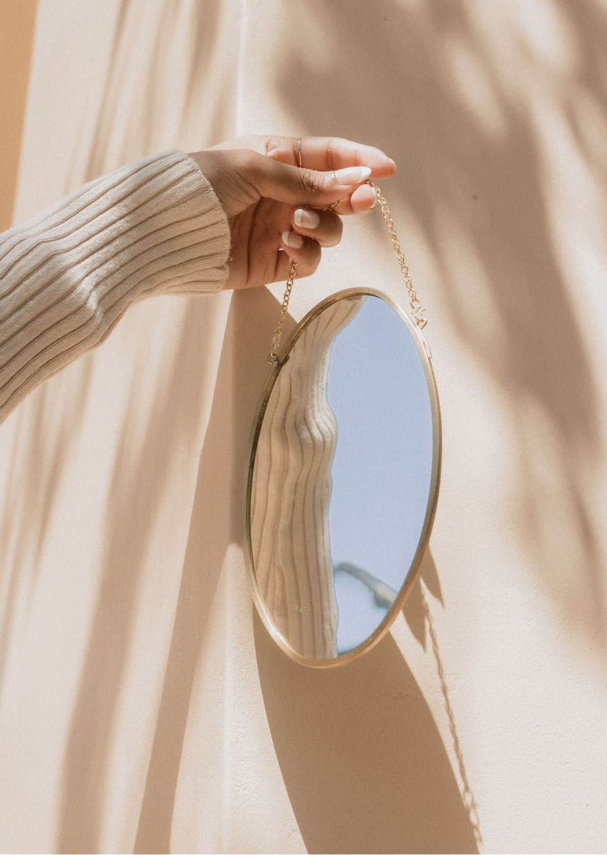 einen Frauenhand hält einen ovalen Spiegel an einer goldenen Kette. Alles ist in gebrochenem Weiß gehalten