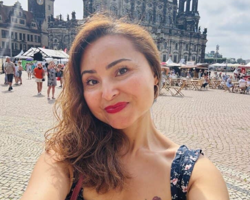 So sieht 46 aus. Eine Frau macht ein Selfie auf einem sonnigen Platz vor einem historischen Gebäude mit Türmen und barocker Architektur. Sie lächelt, trägt ein dunkles, geblümtes Kleid und roten Lippenstift. Im Hintergrund sind Menschen, Tische und Marktstände zu sehen.