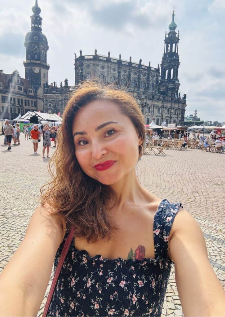Eine Frau macht ein Selfie auf einem sonnigen Platz vor einem historischen Gebäude mit Türmen und barocker Architektur. Sie lächelt, trägt ein dunkles, geblümtes Kleid und roten Lippenstift. Im Hintergrund sind Menschen, Tische und Marktstände zu sehen.