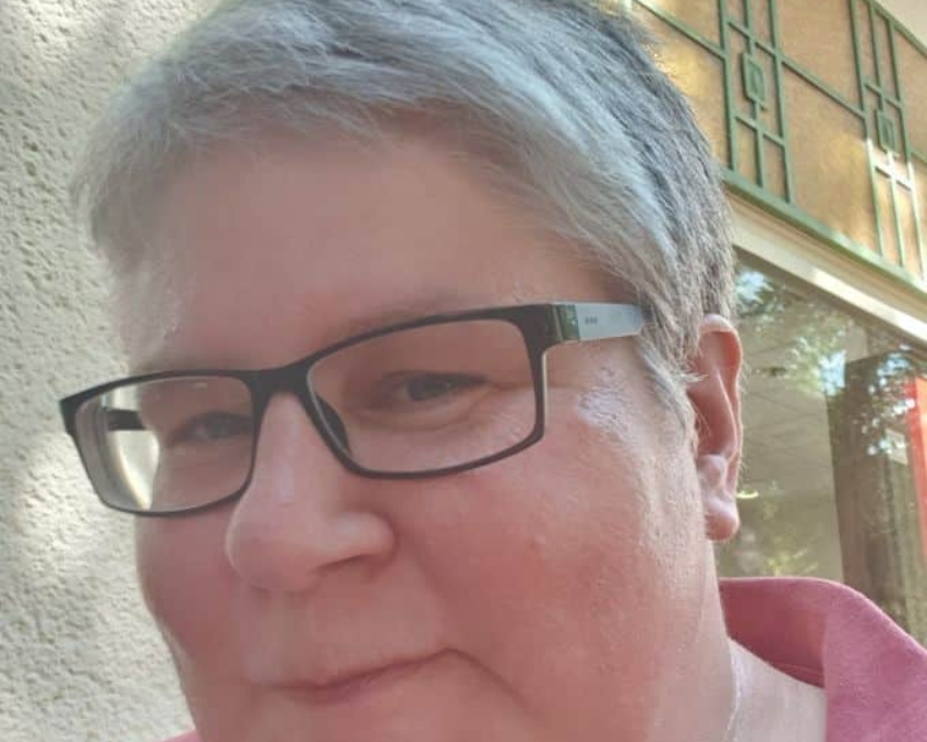 Frau mit Kurzhaarschnitt und grauen Haaren. Sie tr&auml;gt eine schwarze Brille und schaut zufrieden aus.