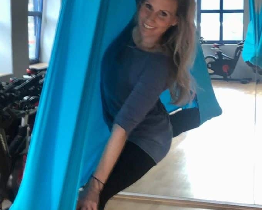 So sieht 47 aus. Frau mit macht Yoga in blauem Tuch. Sie trägt einen langen blonden Pferdeschwanz und freut sich sehr.