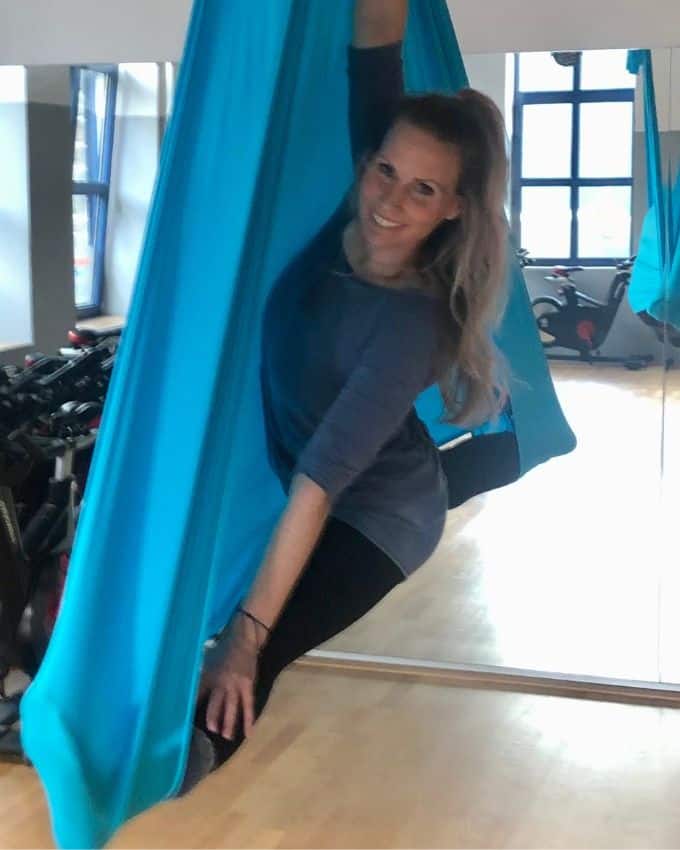 Frau mit macht Yoga in blauem Tuch. Sie trägt einen langen blonden Pferdeschwanz und freut sich sehr.