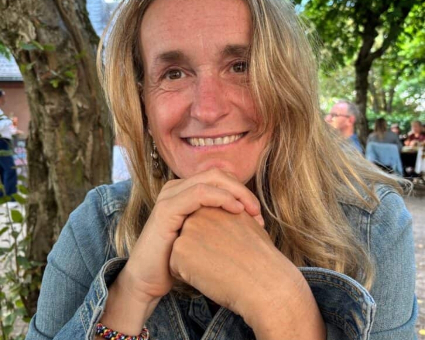 So sieht 54 aus. Eine Frau sitzt im Freien unter Bäumen und lächelt in die Kamera. Sie stützt ihr Kinn auf ihre Hände, trägt eine Jeansjacke und ein buntes Armband. Im Hintergrund sind grüne Blätter, Baumstämme und unscharf weitere Personen in einem Café- oder Gartenbereich zu sehen.