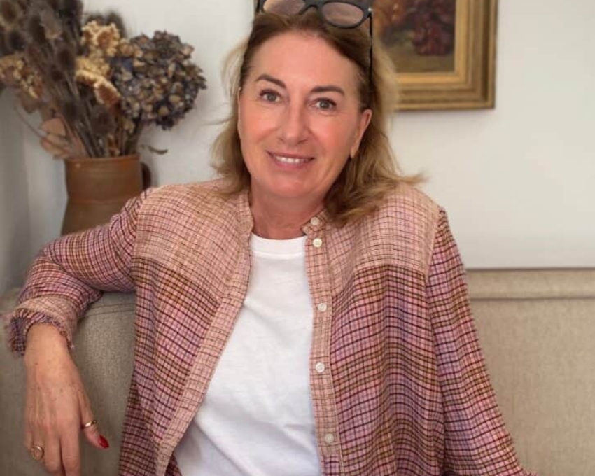 So sieht 61 aus. Eine Frau sitzt entspannt auf einem Sofa in einem gemütlichen Innenraum. Sie lächelt in die Kamera, trägt ein weißes T-Shirt und eine rosafarbene, gemusterte Bluse, und hat eine Brille auf dem Kopf. Im Hintergrund hängen ein gerahmtes Bild und eine Wandlampe, daneben steht eine Vase mit getrockneten Blumen.