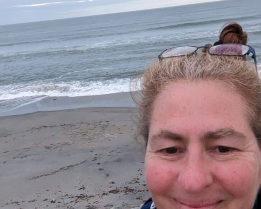 So sieht 60 aus. Eine Frau macht ein Selfie am Strand. Im Hintergrund sind das Meer mit sanften Wellen, ein bewölkter Himmel und ein sandiger Strand zu sehen. Sie lächelt leicht, trägt ihre Haare hochgebunden und hat eine Sonnenbrille auf dem Kopf.