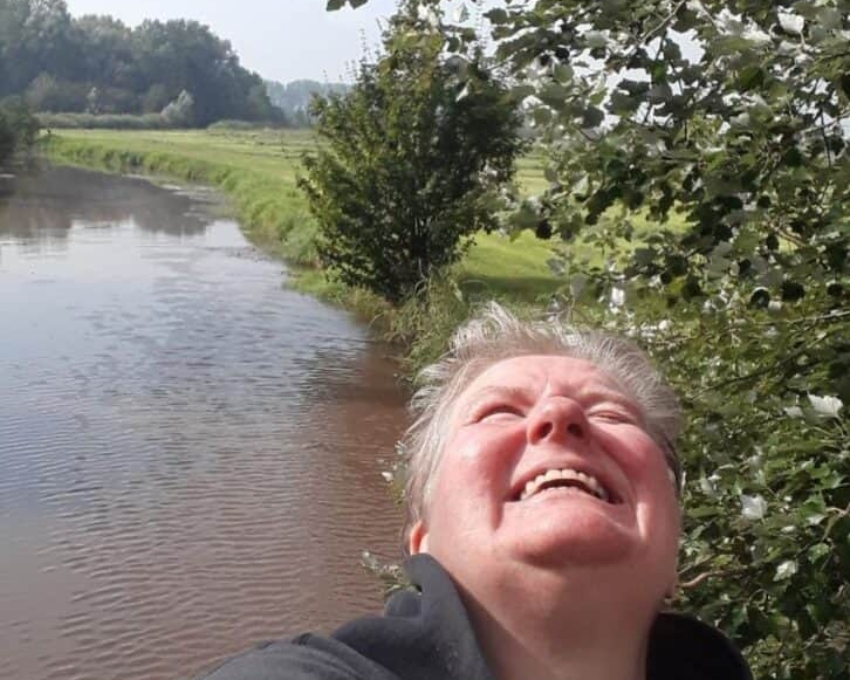 So sieht 60 aus. Eine Person macht ein Selfie im Freien an einem Fluss. Sie schaut lachend nach oben, trägt einen dunklen Hoodie, und im Hintergrund sind ein ruhiger Wasserlauf, grüne Wiesen und Bäume zu sehen.