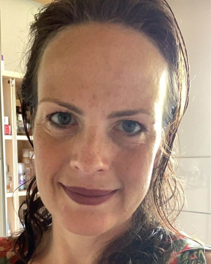 Selfie einer 47-jährigen Frau. Die braunen Haare gehen glatt nach hinten. Sie schaut zufrieden in die Kamera, trägt einen matt-dunkelroten Lippenstift. Sie steht im Badezimmer.