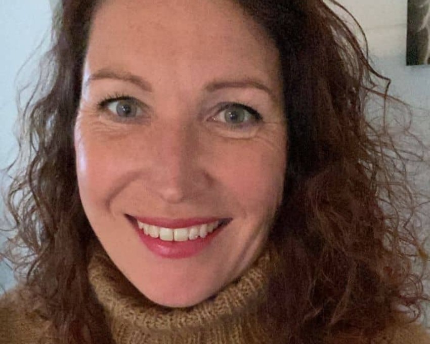 So sieht 48 aus. 48 jährige Frau mit Sommersprossen und rotem Lippenstift und lockigen langen Haaren, schaut zufrieden in die Kamera. Sie trägt einen hellbraunen Rollkragenpullover.