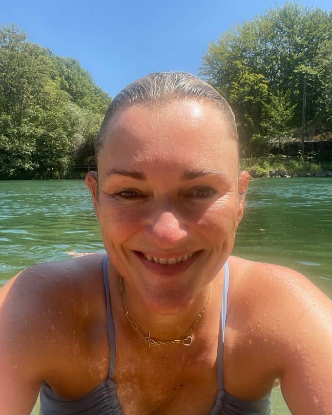 49 jährige Frau schaut mit dem Oberkörper aus dem Wasser. Sue lächelt in die Kamera und trägt ein hellblaues Bikini Oberteil.