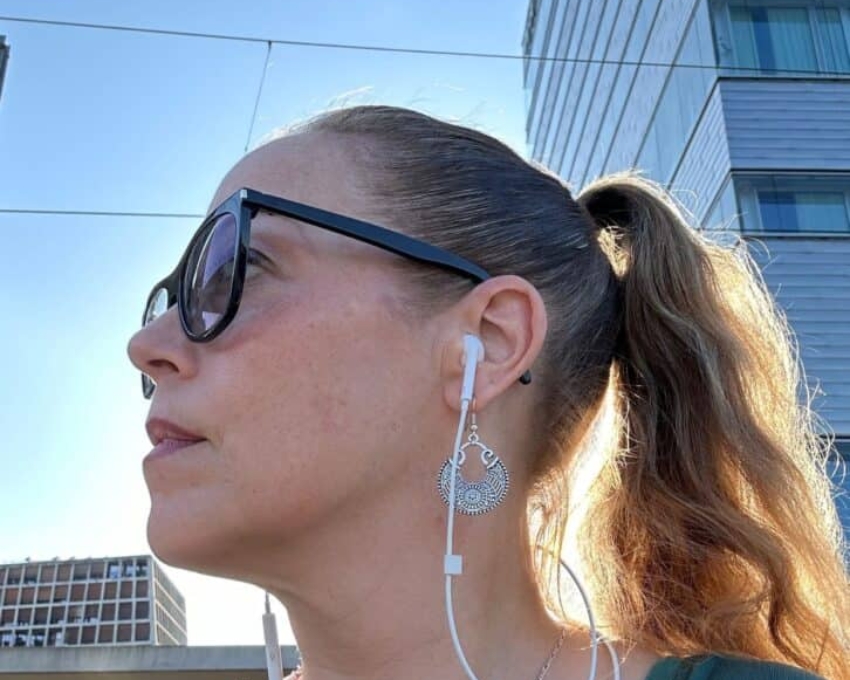 So sieht 49 aus. Frau mit Sonnenbrille und Ohrhörern, im Profil bei Tageslicht, mit zurückgebundenen Haaren und auffälligem Ohrring, vor moderner städtischer Architektur.