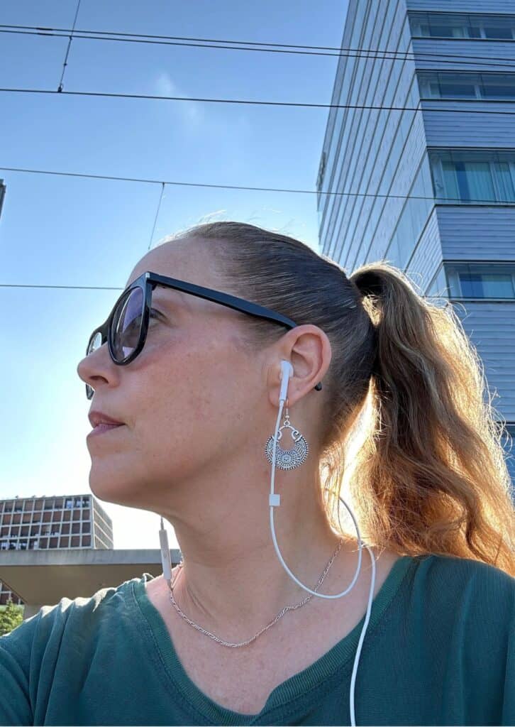 Frau mit Sonnenbrille und Ohrhörern, im Profil bei Tageslicht, mit zurückgebundenen Haaren und auffälligem Ohrring, vor moderner städtischer Architektur.
