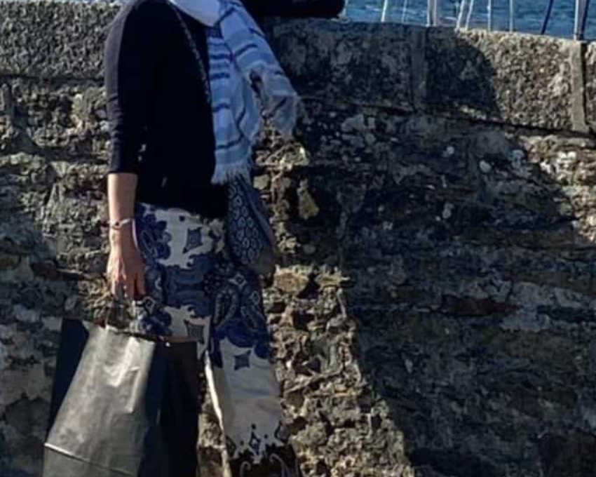 So sieht 51 aus. Eine Frau steht an der mauer zu einem Segelhafen. Sie trägt blonde kurze Haare, eine Brille und eine schwarze Papiertasche in der Hand. Sie trägt einen auffällig großen weißen Schal um den Hals.