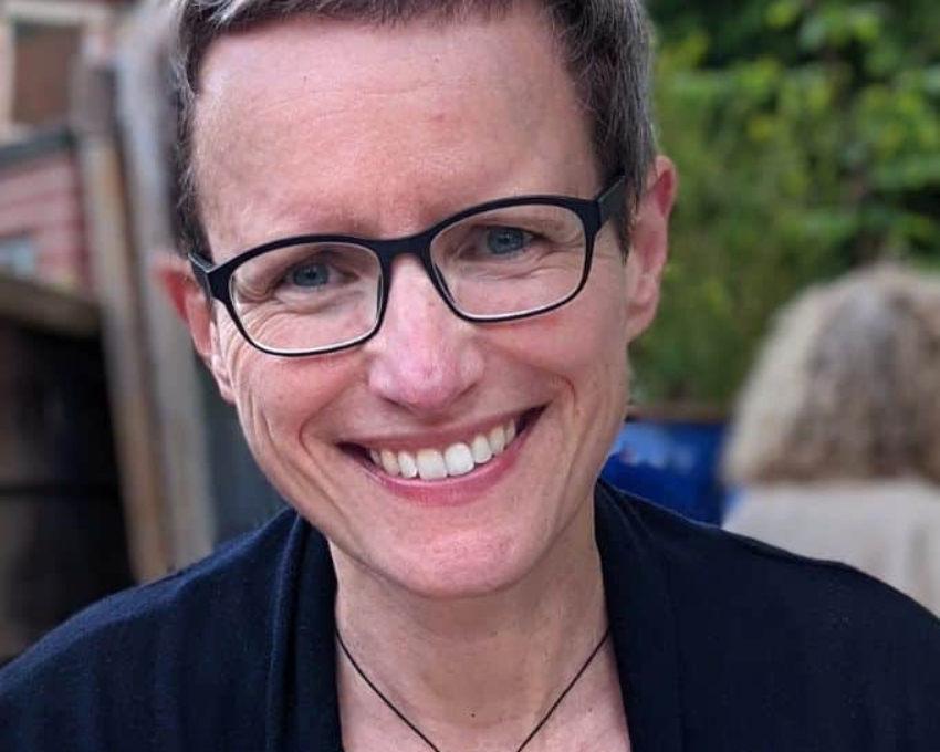 So sieht 52 aus. 52 jährige Frau mit grauem Kurzhaarschnitt und schwarzer eckiger Brille lacht in die Kamera. Sie trägt einen auffälligen goldenen Kettenanhänger an einem schwarzen Band.