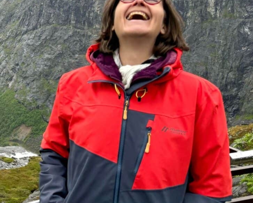 So sieht 53 aus. Lachende Frau in roter und dunkelgrauer Outdoorjacke vor einer felsigen Berglandschaft mit Nebel; sie steht an einem Geländer und wirkt fröhlich und unbeschwert.