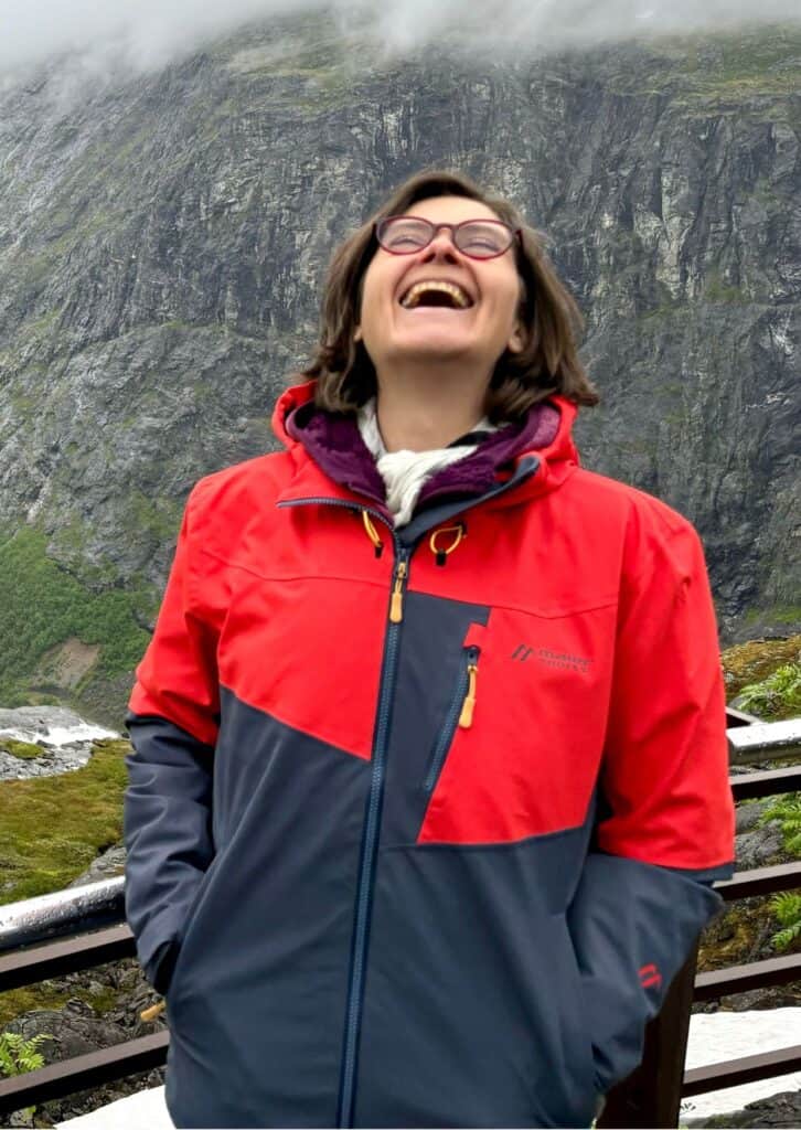 Lachende Frau in roter und dunkelgrauer Outdoorjacke vor einer felsigen Berglandschaft mit Nebel; sie steht an einem Geländer und wirkt fröhlich und unbeschwert.