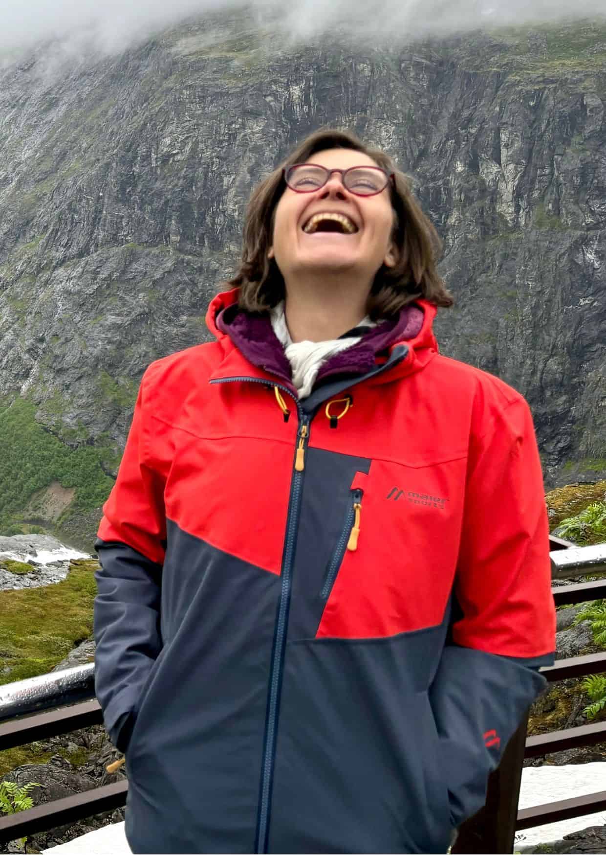 Lachende Frau in roter und dunkelgrauer Outdoorjacke vor einer felsigen Berglandschaft mit Nebel; sie steht an einem Geländer und wirkt fröhlich und unbeschwert.