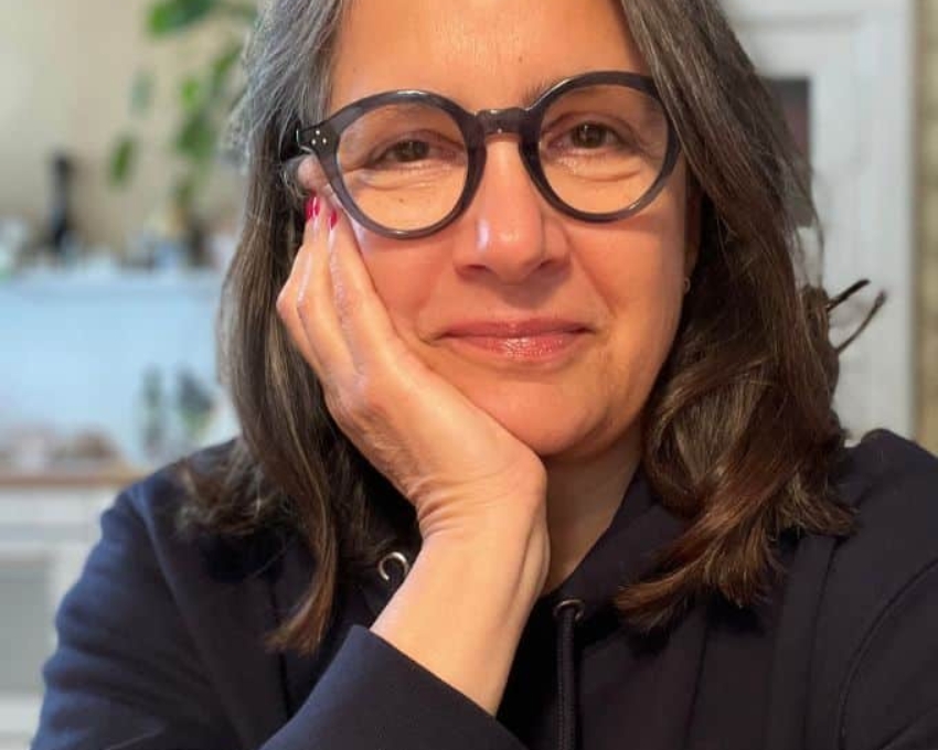 So sieht 57 aus. 57 jährige Frau, die einen dunkelblauen Hoodie mit rotem Print trägt und eine dunkelgraue Brille aufhat, schaut zufrieden in die Kamera.