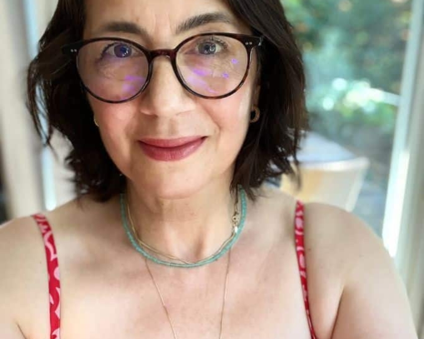 So sieht 59 aus. 59 jährige Frau mit dunkelbraunem Bob und großer schwarzer Brille schaut zuversichtlich in die Kamera. Sie trägt ein rotes Trägerkleid und eine jade farbene Kette um den Hals