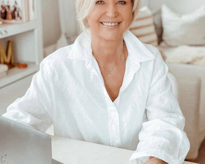 So sieht 60 aus. Sechzigjährige Frau mit weiß-blondem, kurzem Bob sitzt mit weißer Bluse vor dem Laptop und strahlt in die Kamera. Man sieht im Hintergrund ihr in Weiß gehaltenes Wohnzimmer.