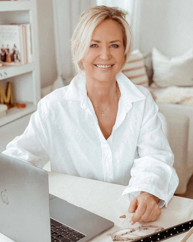 Sechzigjährige Frau mit weiß-blondem, kurzem Bob sitzt mit weißer Bluse vor dem Laptop und strahlt in die Kamera. Man sieht im Hintergrund ihr in Weiß gehaltenes Wohnzimmer.
