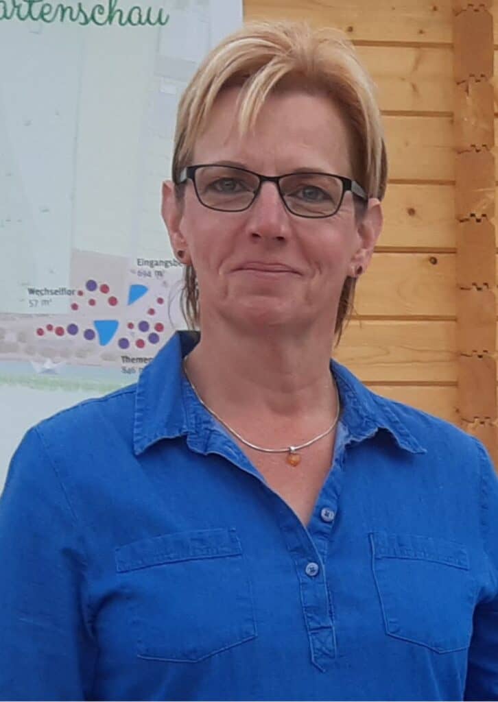 Porträt einer Frau mittleren Alters mit kurzen blonden Haaren, Brille und blauem Hemd, stehend vor einer Holzfassade und einem Plakat.