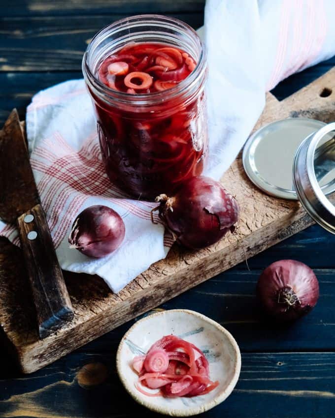 Pickled red onions in a glass container on a wooden cutting board while raw onions surround the container