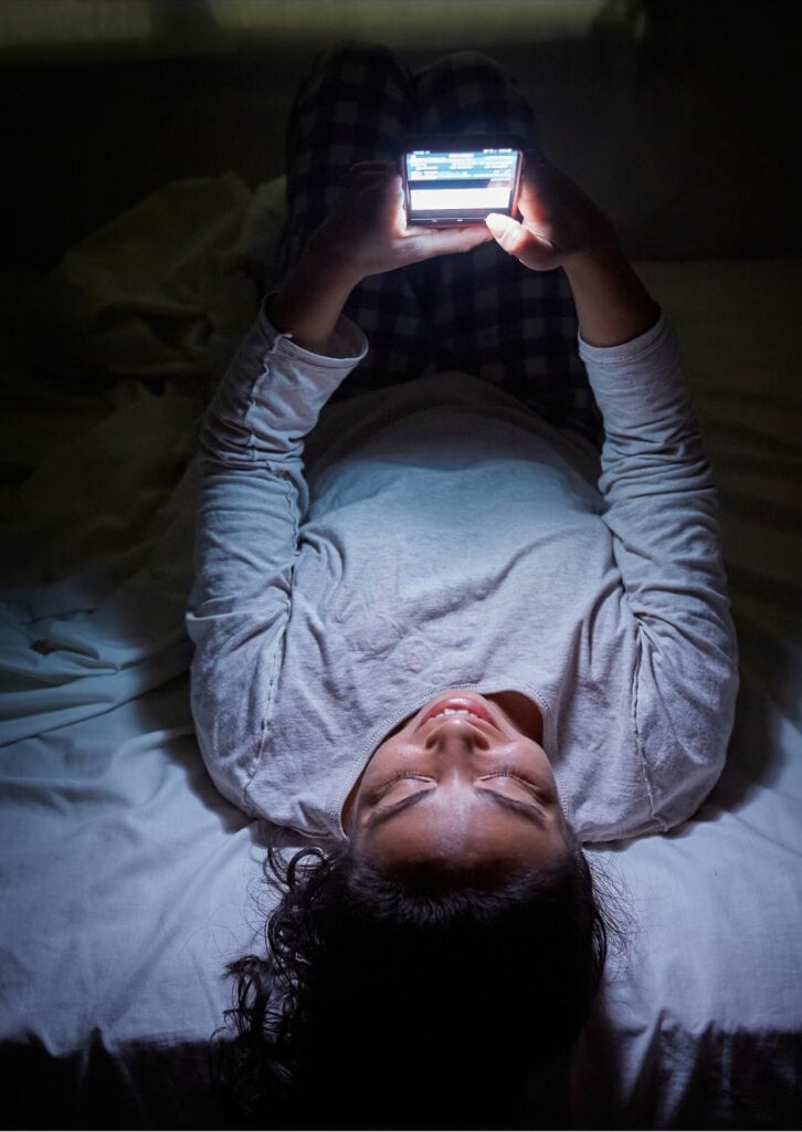eine Frau mit langen schwarzen Haaren, die auf dem Bett liegt und mit dem Handy SMS schreibt
