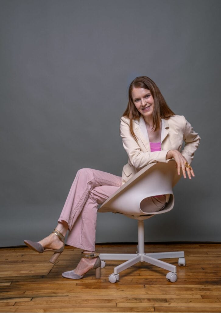 Lächelnde Frau mittleren Alters in modischer rosa Hose und beigem Blazer sitzt entspannt auf einem modernen Bürostuhl vor grauem Hintergrund