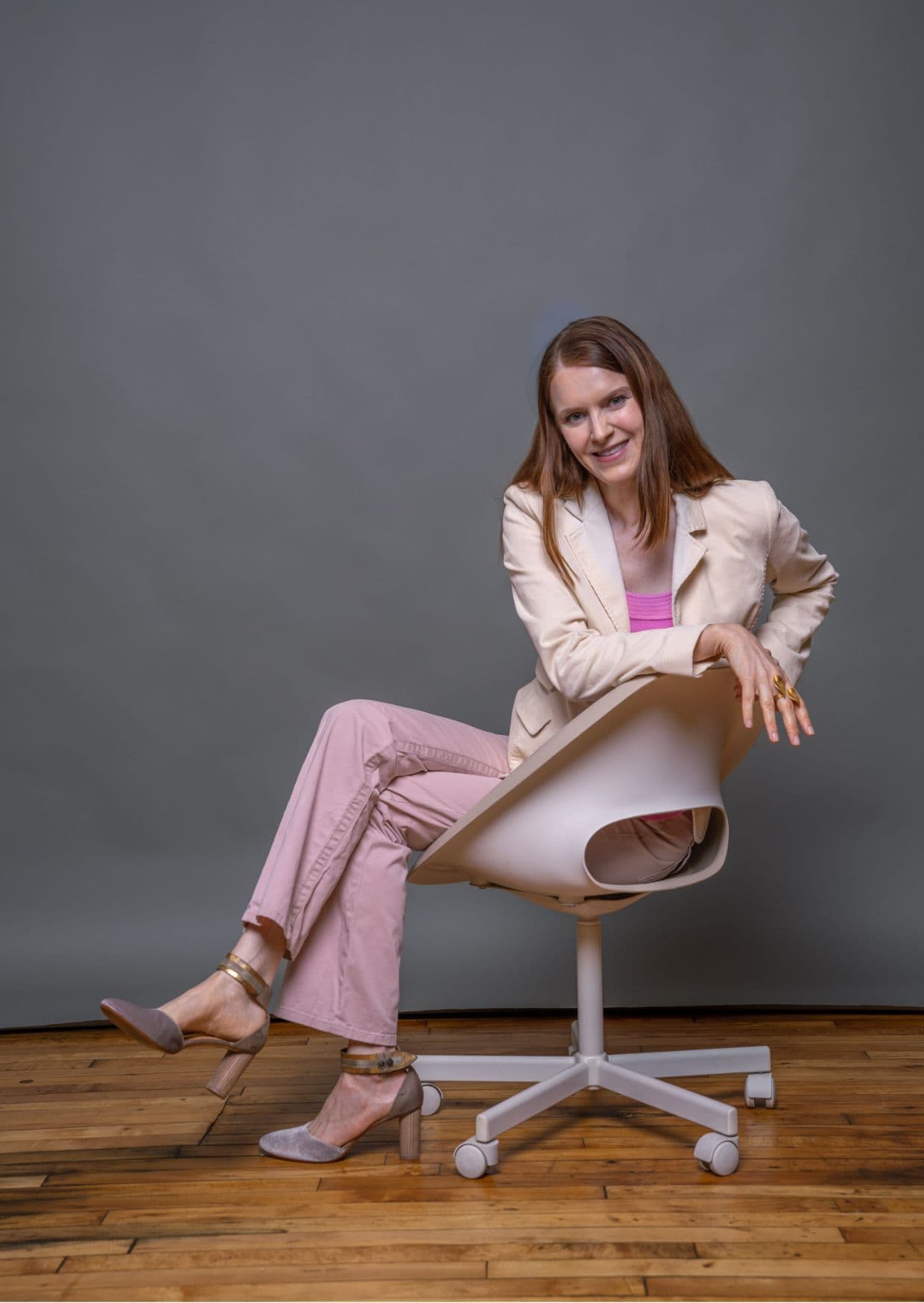 Lächelnde Frau mittleren Alters in modischer rosa Hose und beigem Blazer sitzt entspannt auf einem modernen Bürostuhl vor grauem Hintergrund