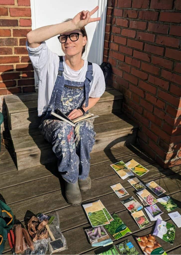 Frau in Maleranzug sitzt auf einer Holzterrasse und plant ihren Garten mit verschiedenen Gemüsesaatguttüten.