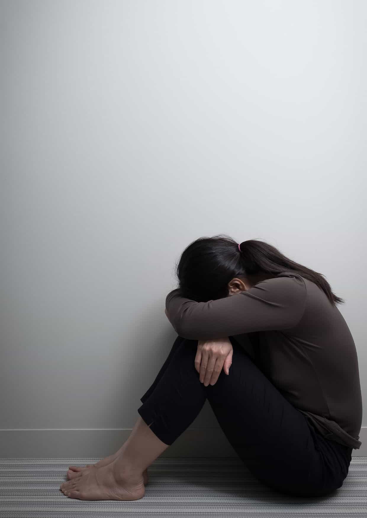 Eine Frau sitzt zusammengesunken auf dem Boden mit dem Rücken zur Wand und dem Kopf auf die Knie gelegt, was Traurigkeit und Niedergeschlagenheit ausdrückt.