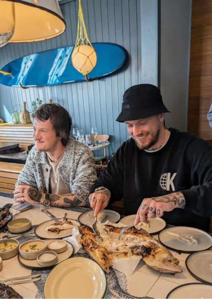Zwei lächelnde Männer sitzen an einem Tisch in einem Restaurant mit maritimem Flair, erkennbar an der blauen Wand und einem Surfbrett als Dekoration. Beide haben sichtbare Tätowierungen und der eine trägt eine schwarze Mütze. Vor ihnen auf dem Tisch liegt ein gegrillter Fisch, an dem einer der Männer gerade schneidet, umgeben von Tellern und Saucenschälchen, bereit zum gemeinsamen Essen.